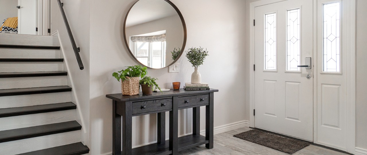 Wide entryway with white walls and white front door with a round mirror hanging above black wooden entrway table.